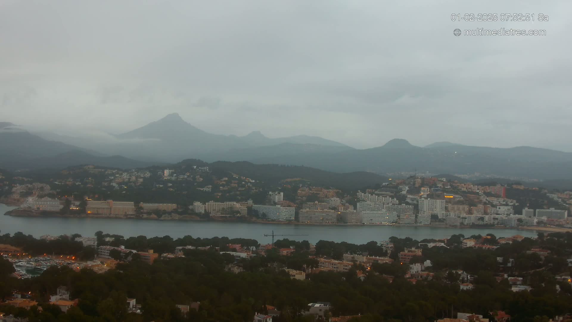 A coastal town nestled among dark green hills and mountains under a cloudy, overcast sky.