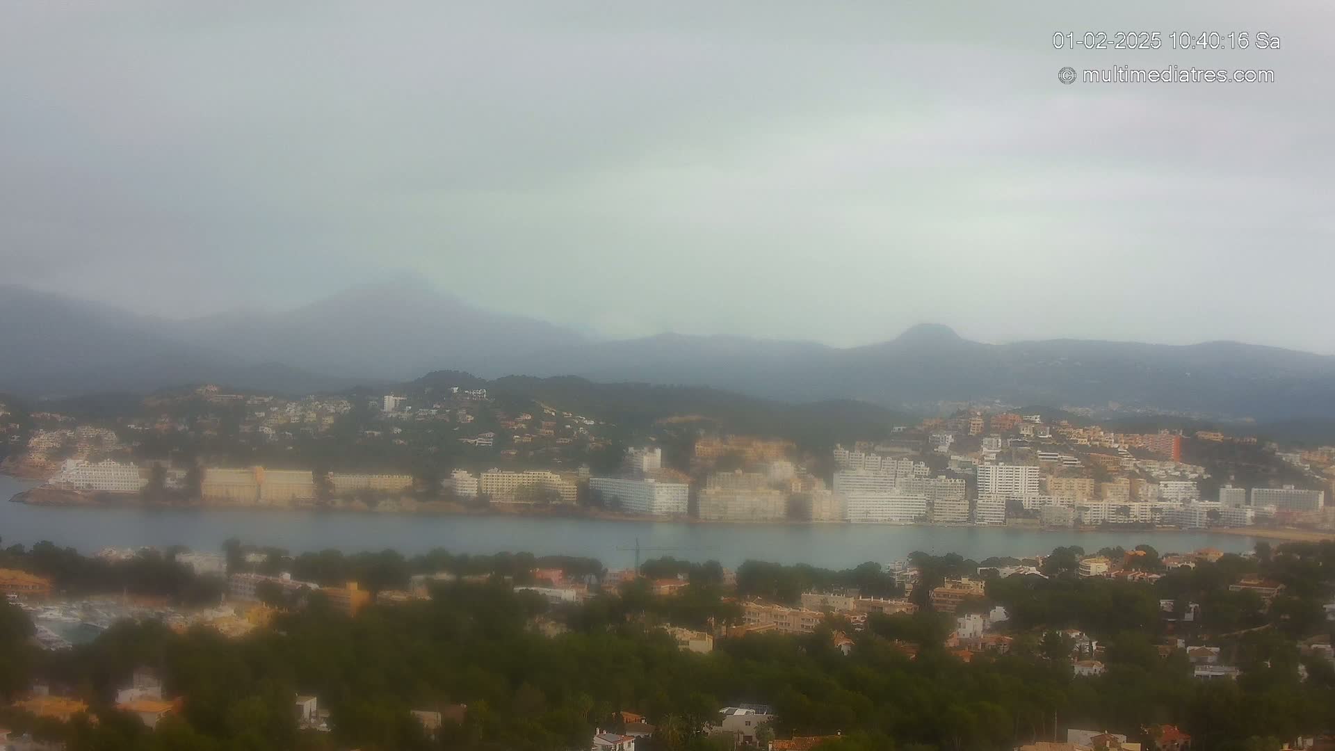 A hazy coastal town with numerous buildings is nestled between a body of water and a range of mountains under an overcast sky.