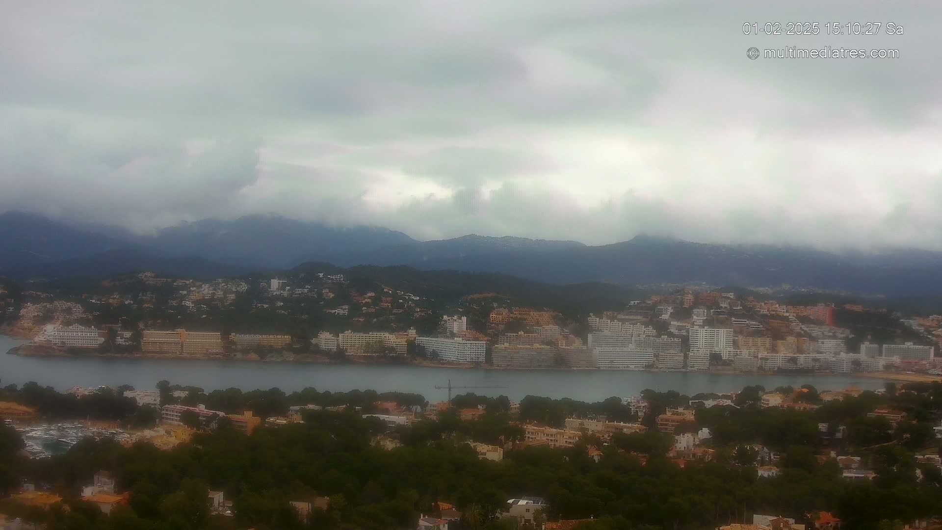 A coastal town nestled among hills and mountains under a cloudy sky, with a body of water separating the town from a larger area of buildings.