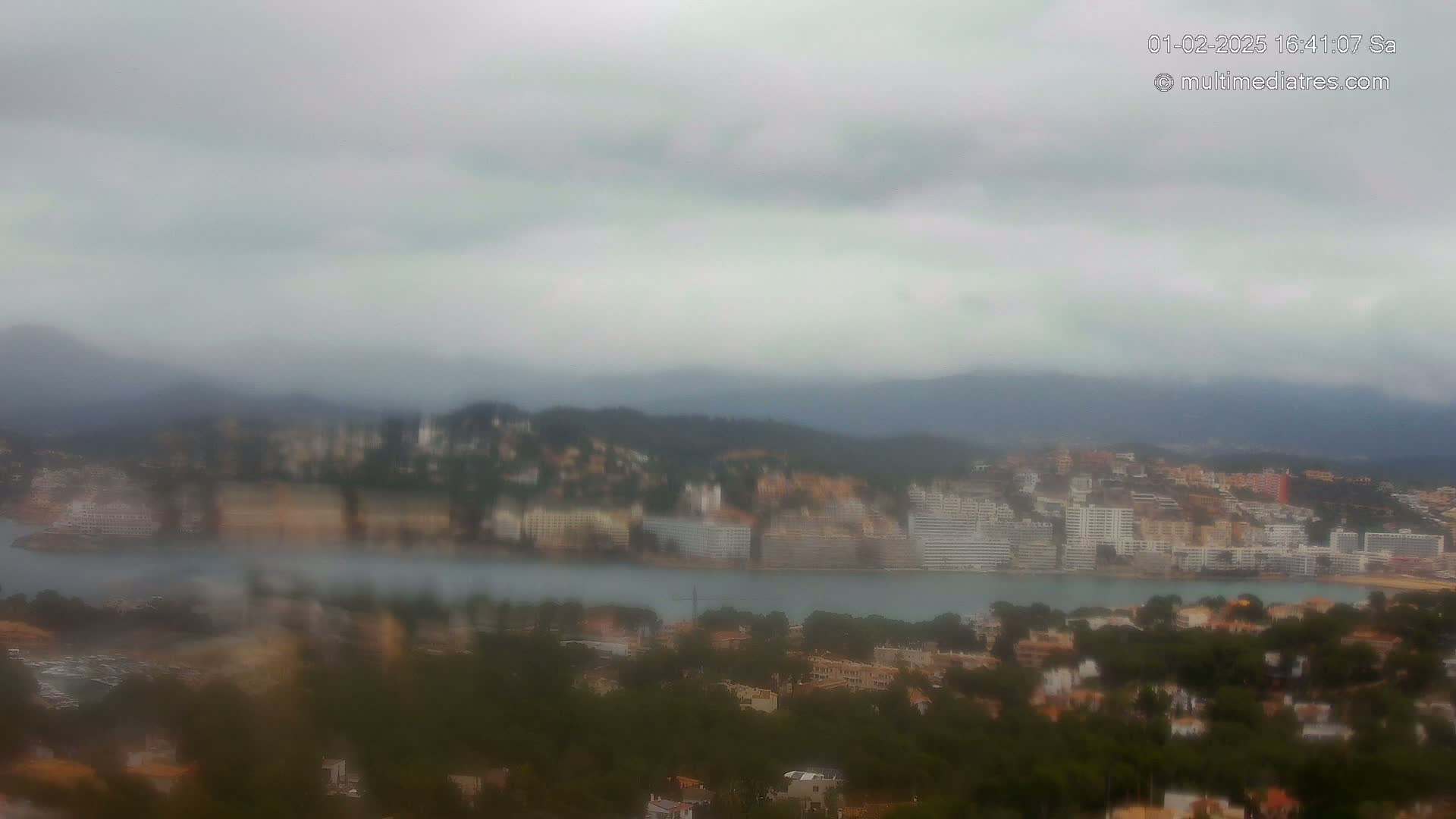 A blurry, rain-obscured view shows a coastal town nestled between hills and a body of water under an overcast sky.