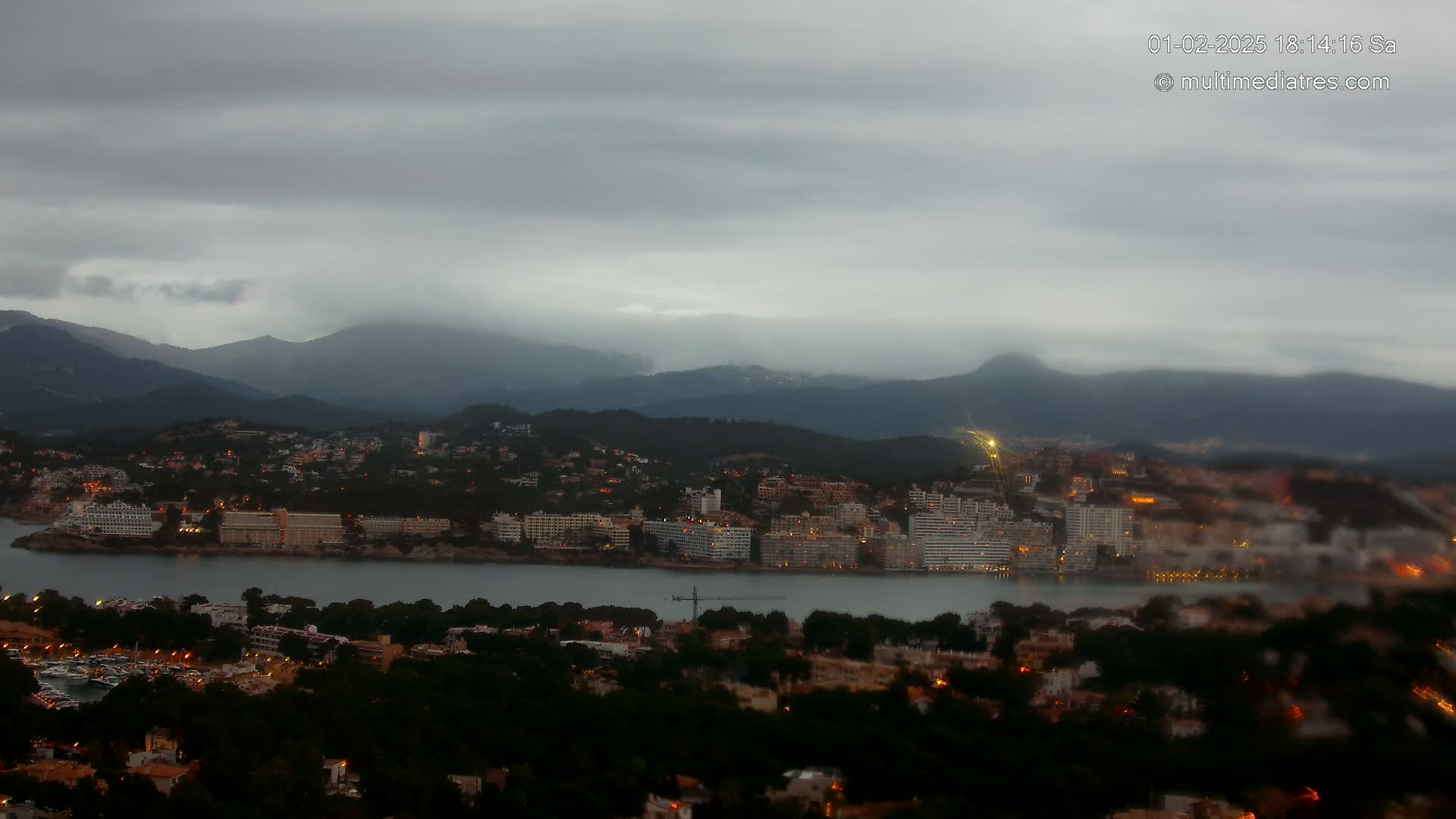 A coastal town with dimly lit buildings and a marina nestles between dark green hills under a cloudy, overcast sky.