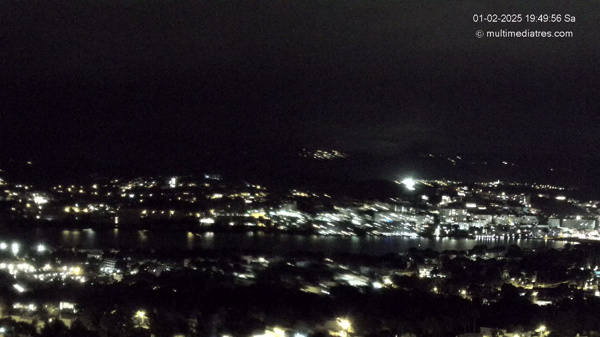 A nighttime, high-angle view of a city situated on a body of water, with its lights illuminating the surrounding hills and dark sky.