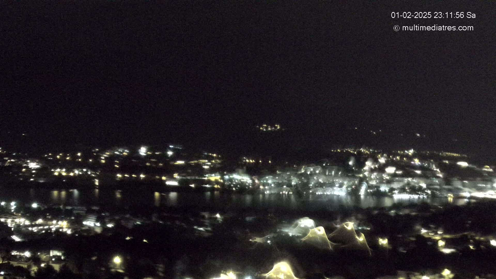 A nighttime, high-angle view shows a city with lights across a body of water, under a clear, dark sky.