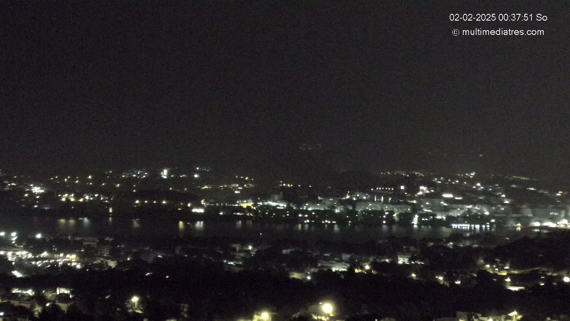 A nighttime, long-range view of a city and body of water, sparsely lit by scattered streetlights.