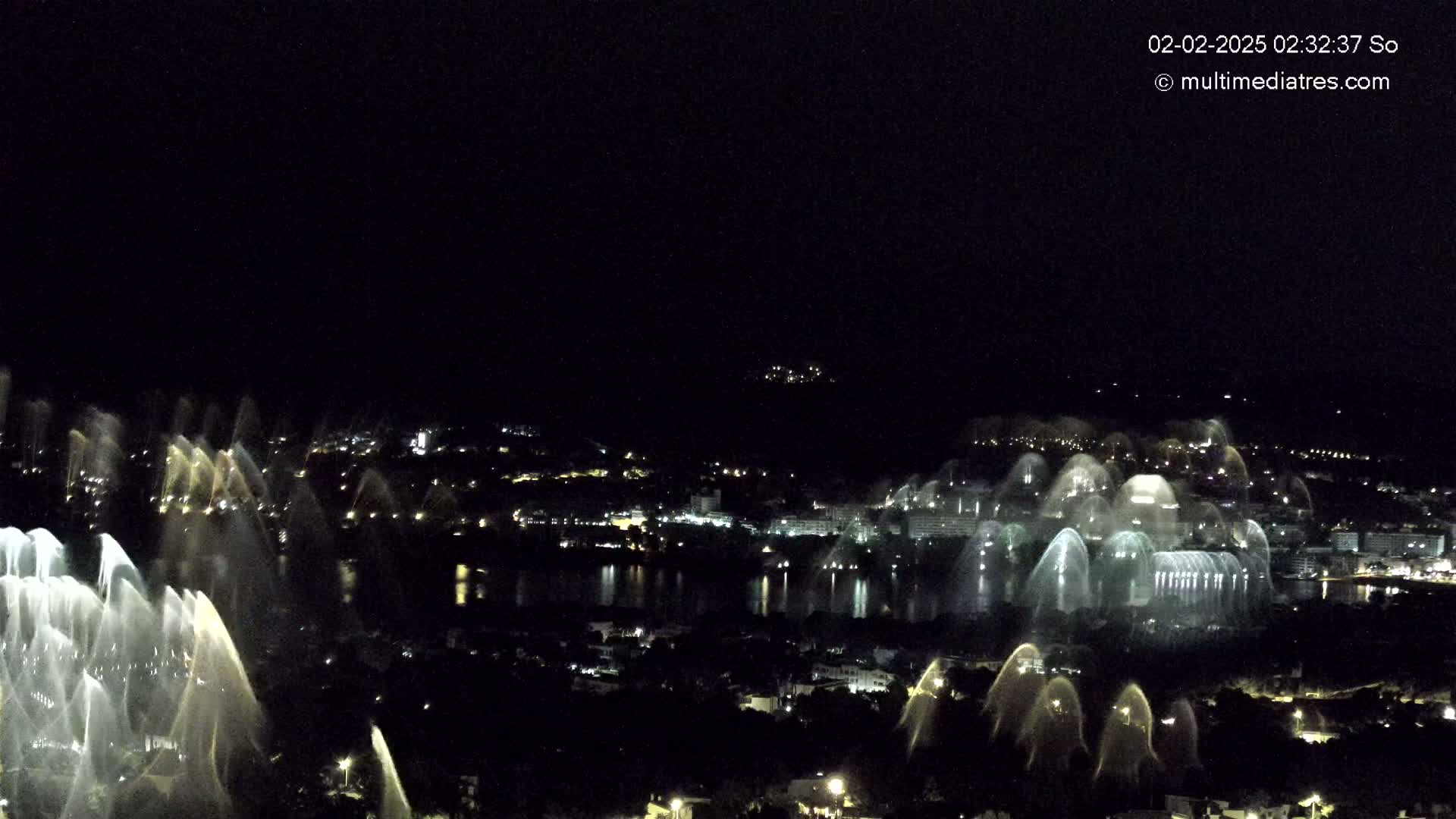 A nighttime view of a city by a body of water, featuring numerous illuminated water fountains.