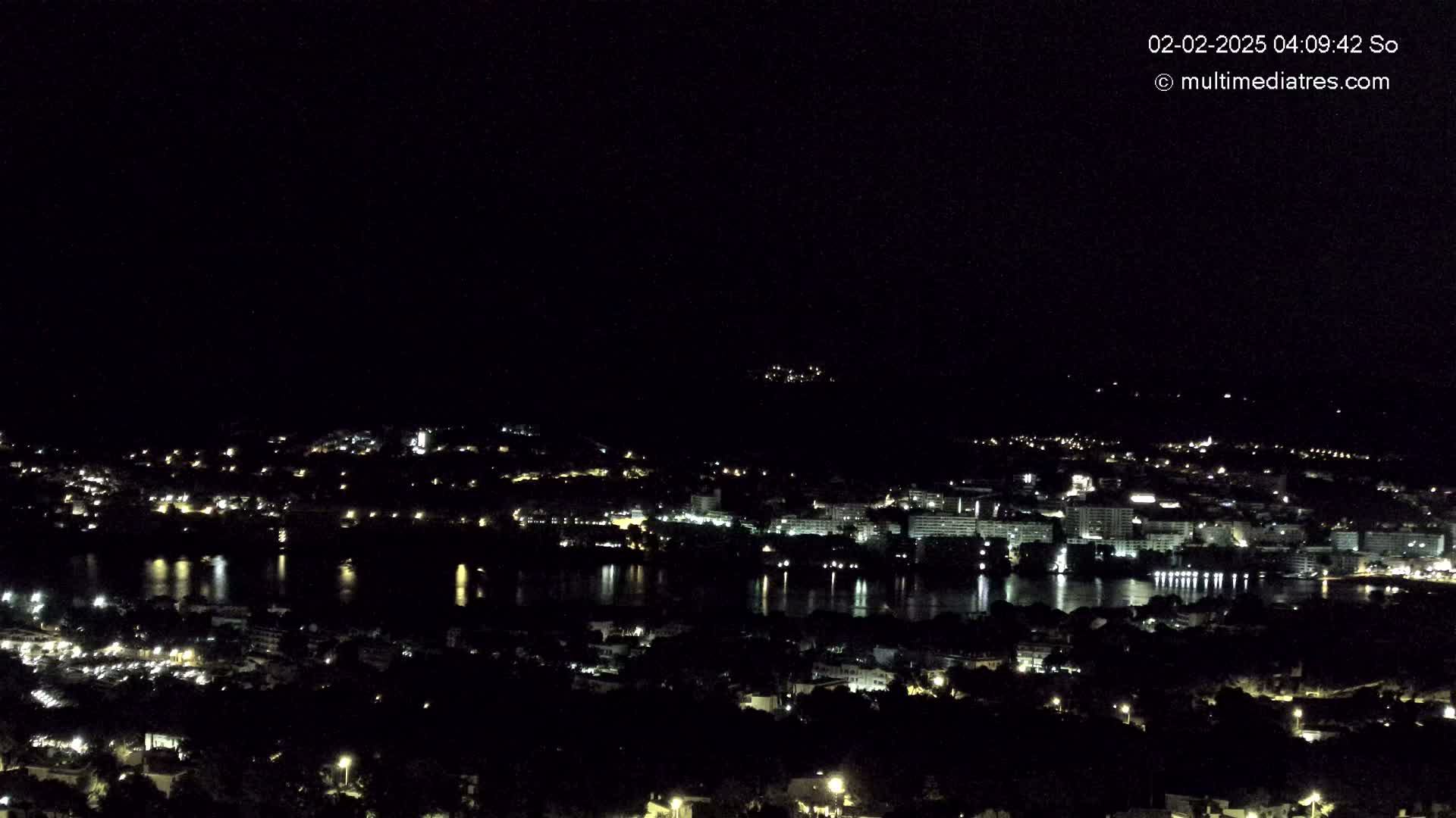A nighttime view of a coastal city with a body of water reflecting city lights under a clear, dark sky.