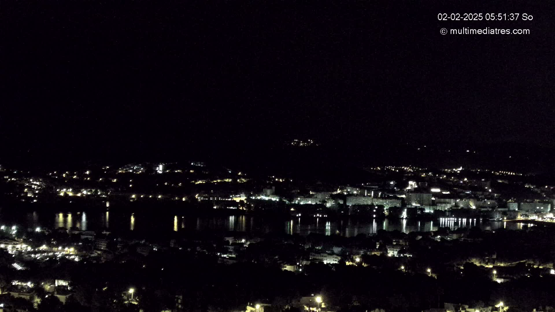 A nighttime, high-angle view of a coastal town with its lights reflecting on a calm body of water under a clear, dark sky.