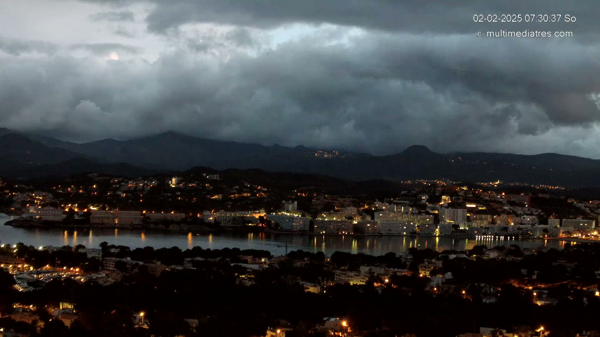 A nighttime coastal city with scattered lights, nestled between dark mountains under a heavily overcast sky.