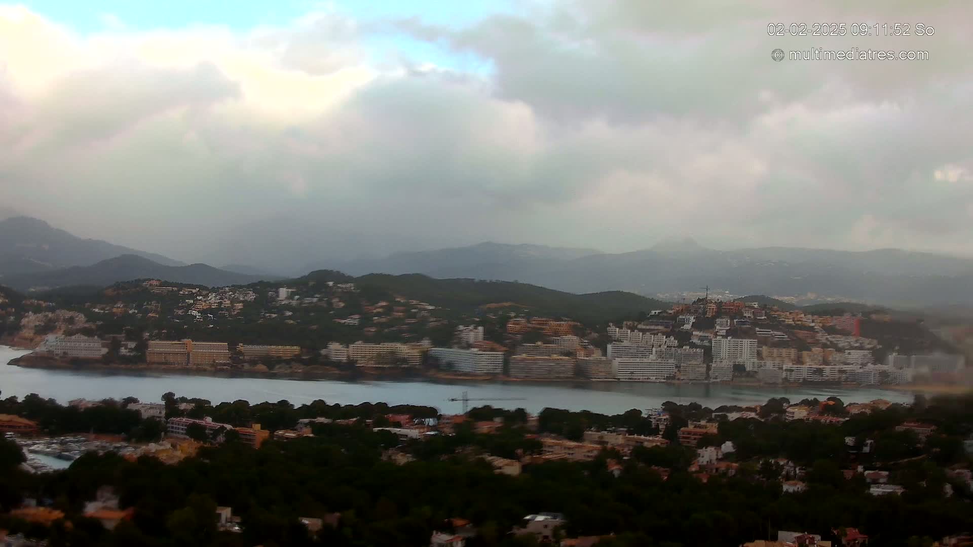 A coastal town nestled in a bay, surrounded by hills under a cloudy sky.