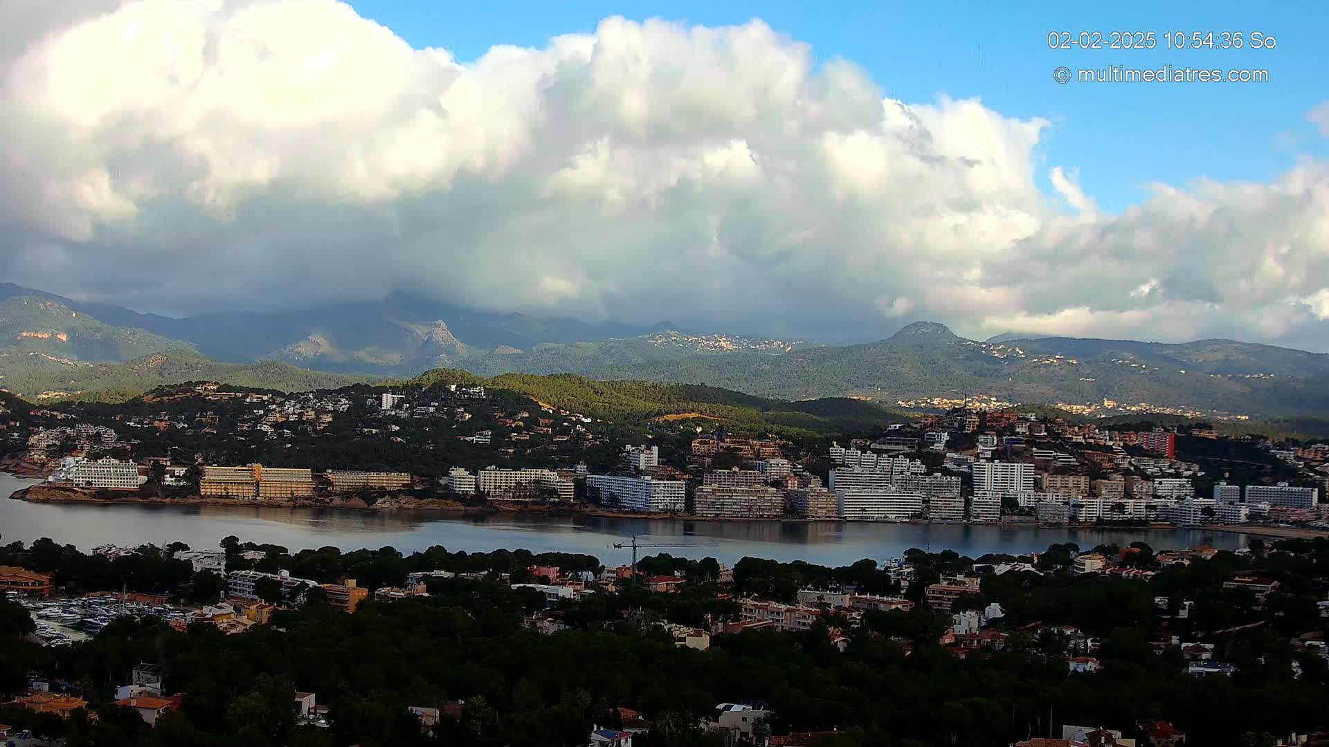 A coastal town nestled between a calm bay and mountains under a partly cloudy sky.