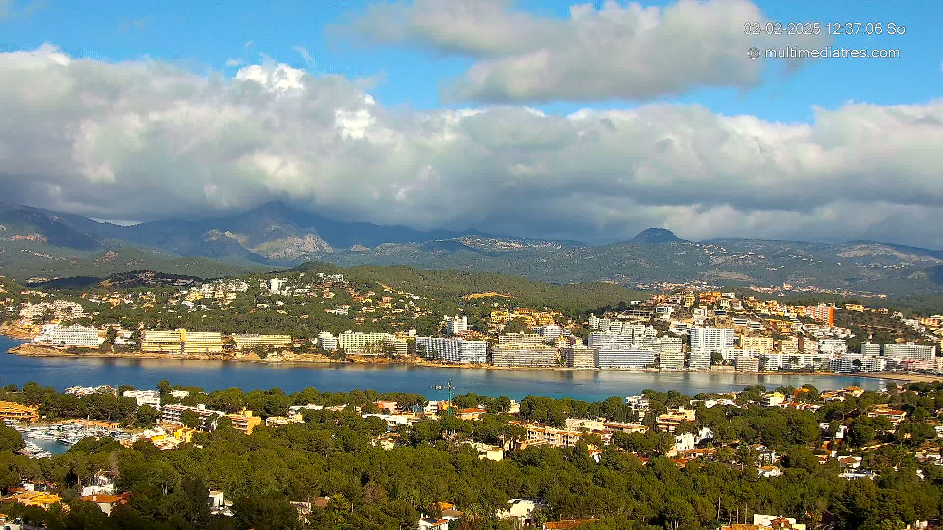 A coastal town with numerous buildings nestled amongst green trees and hills, backed by mountains under a partly cloudy sky.