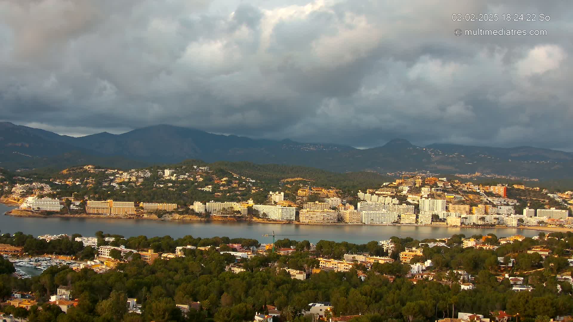 A coastal town nestled amongst green hills and mountains under a cloudy sky.
