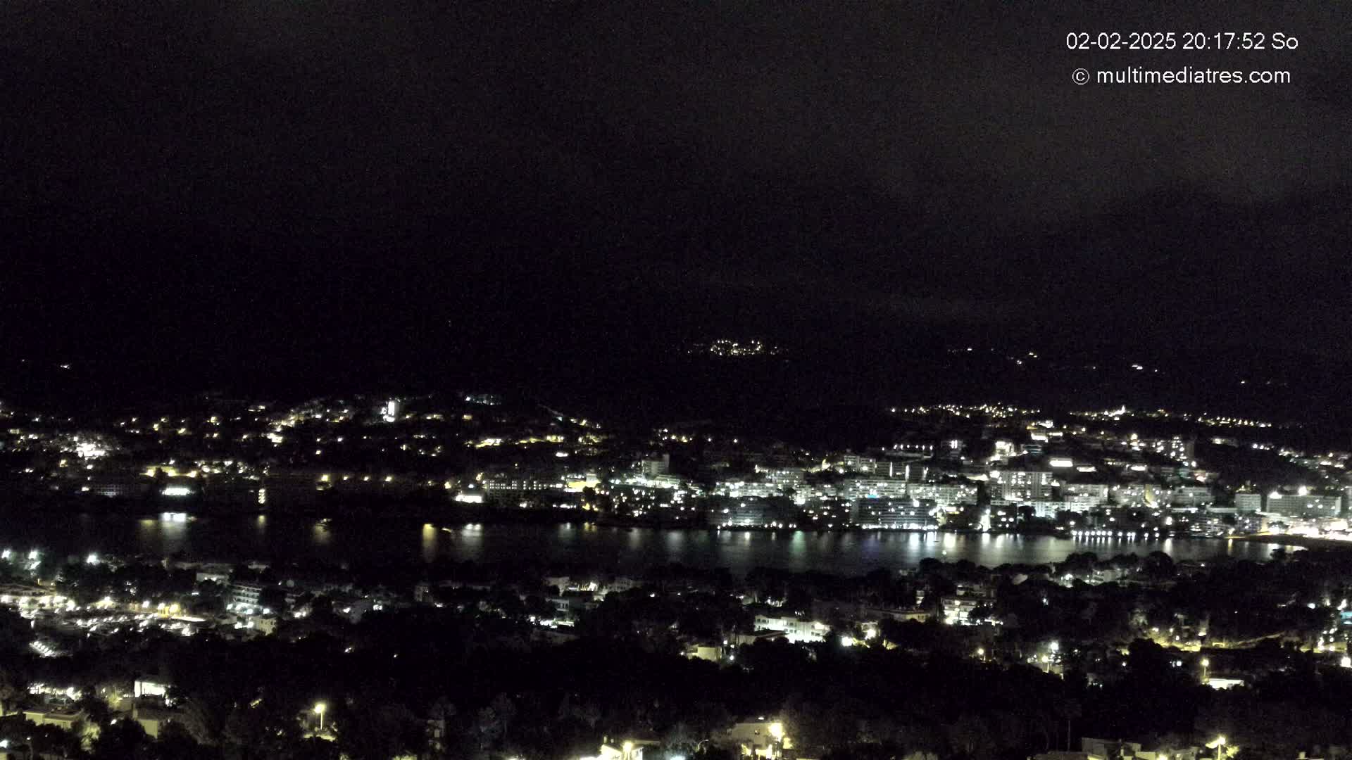 A nighttime, high-angle view shows a city illuminated with lights next to a dark body of water, all under a dark sky.