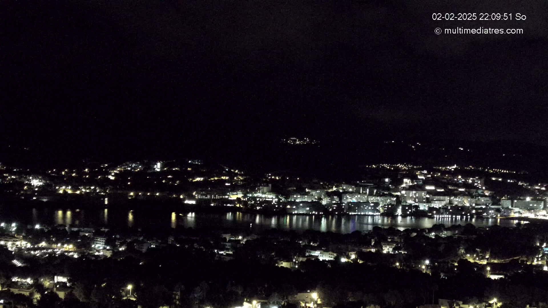 A nighttime, high-angle view of a city illuminated by lights, situated beside a body of water reflecting the city lights.