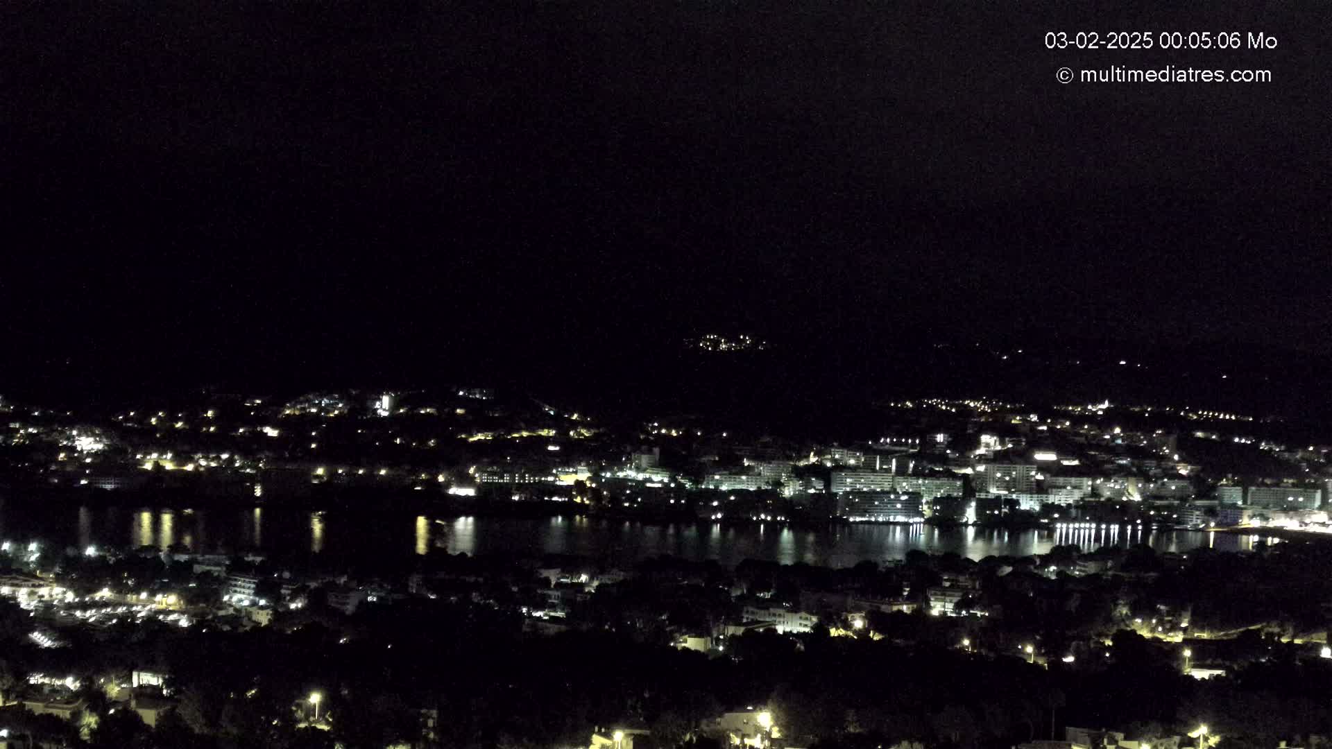 A nighttime view of a coastal town with a body of water reflecting the lights, under a clear, dark sky.