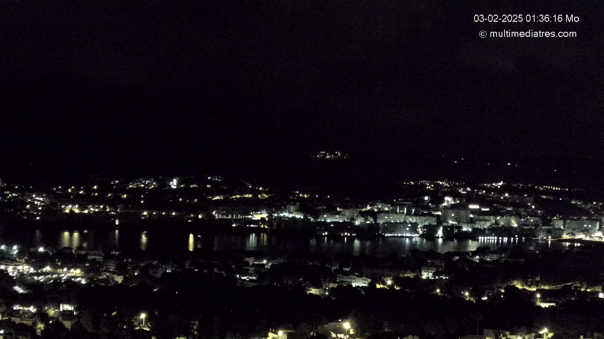A nighttime, high-angle view shows a coastal city illuminated with many small lights surrounding a dark body of water.