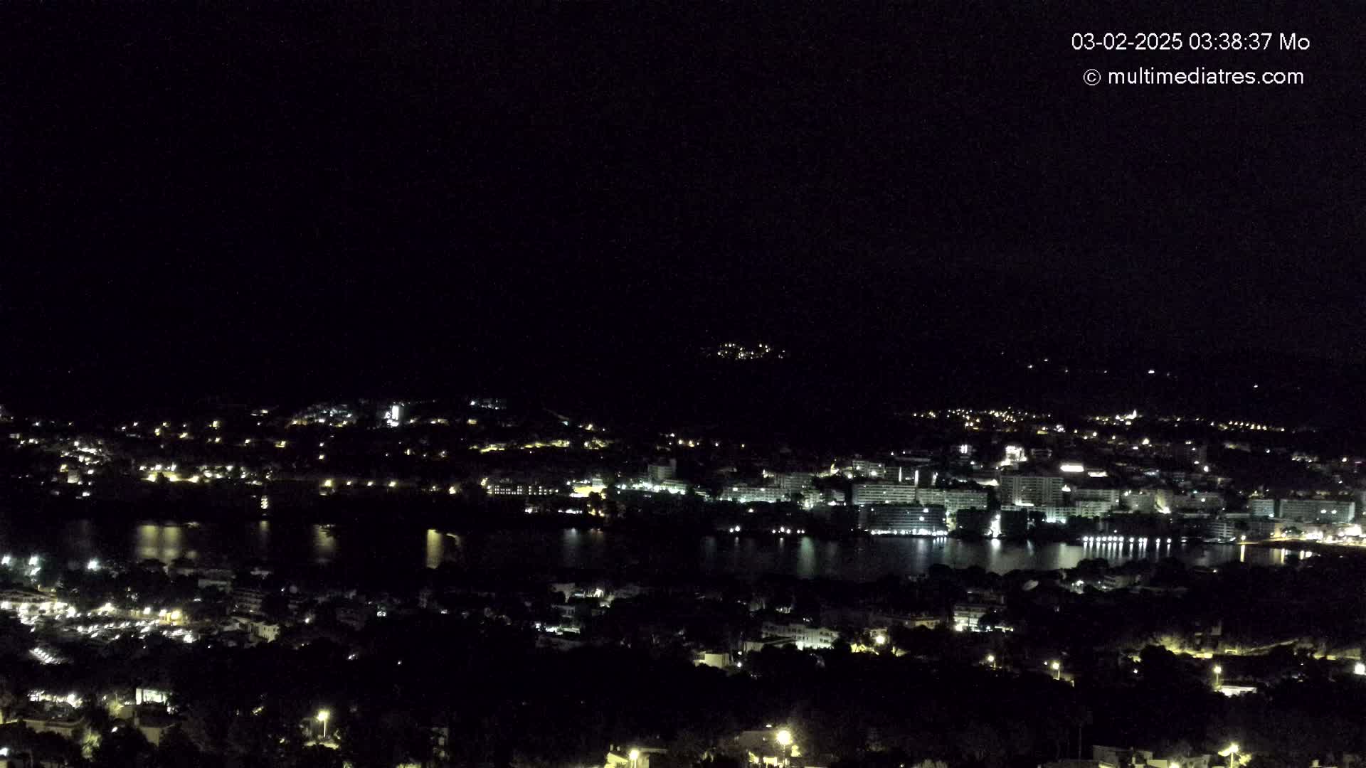 A nighttime aerial view shows a coastal town with its lights illuminated, a body of water reflecting the lights, and dark hills in the background under a clear, dark sky.