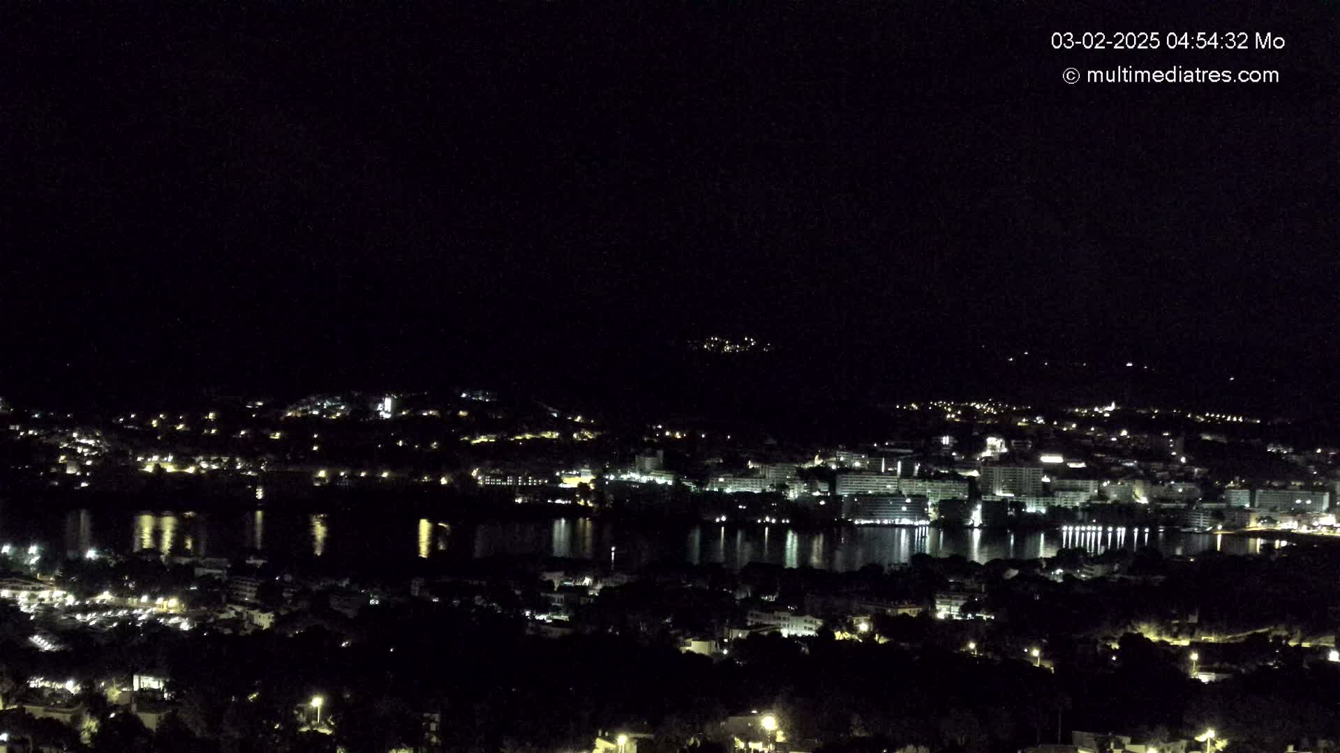 A nighttime, high-angle view shows a coastal town illuminated with lights, reflecting on a calm body of water under a clear, dark sky.