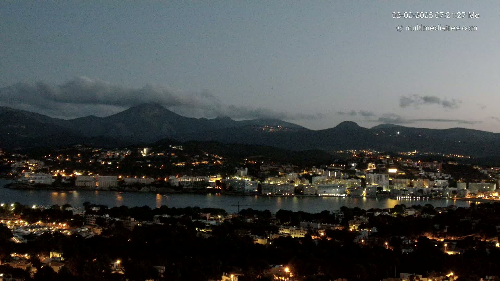 A nighttime view of a coastal city illuminated by lights, nestled beside a calm body of water and backed by dark mountains under a cloudy twilight sky.
