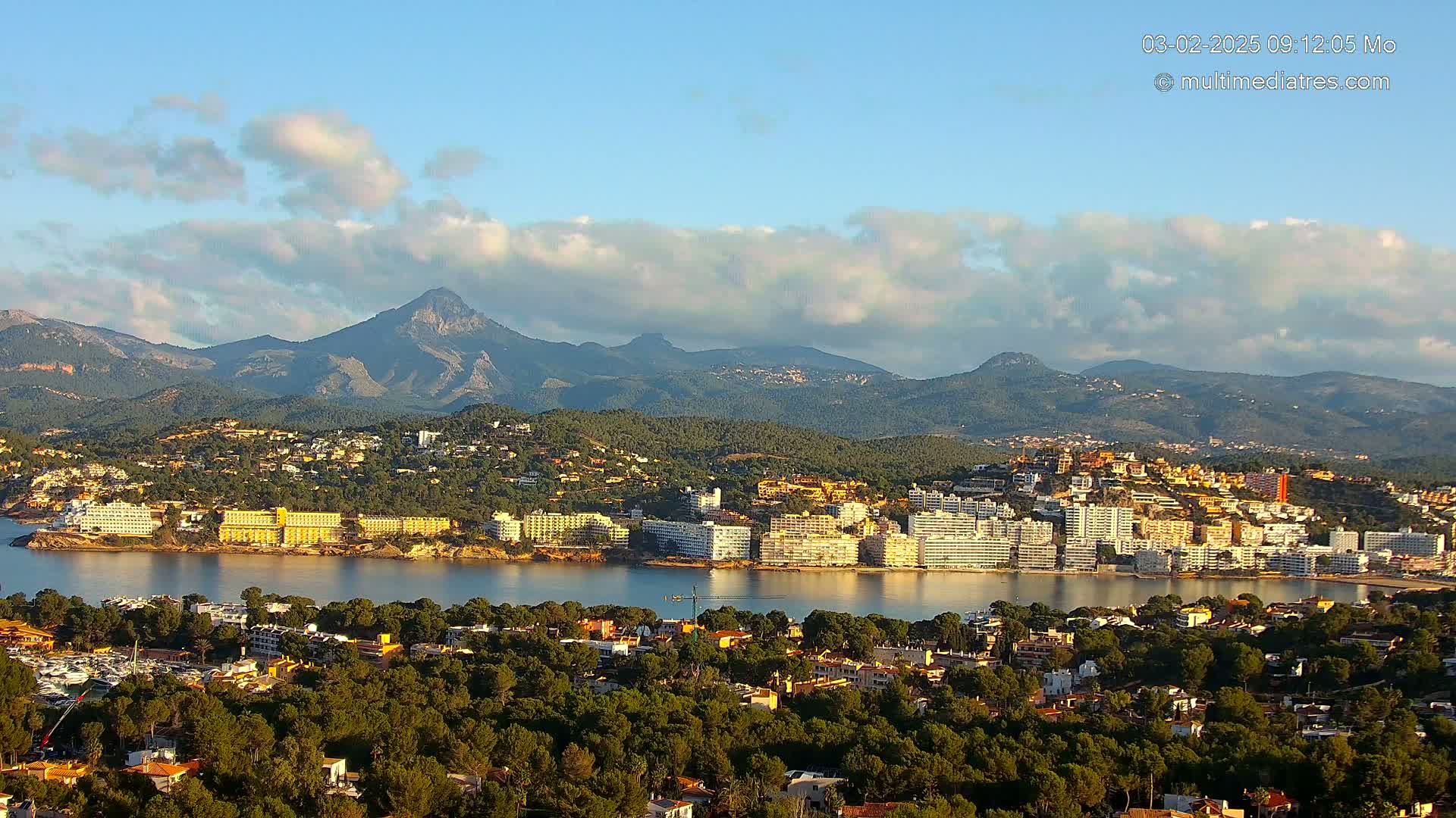 A coastal town nestled at the base of mountains under a partly cloudy sky sits on a bay with many buildings visible.