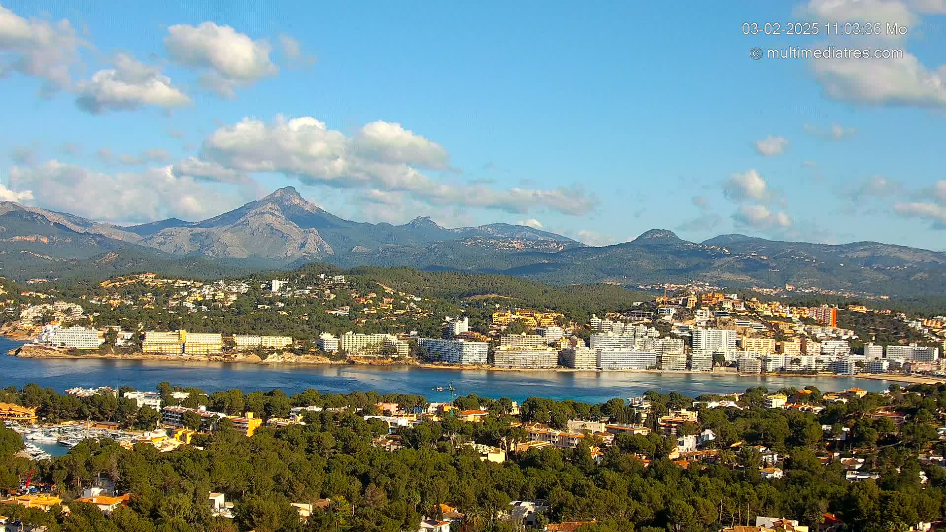 A coastal town nestled among green hills and mountains under a partly cloudy sky.