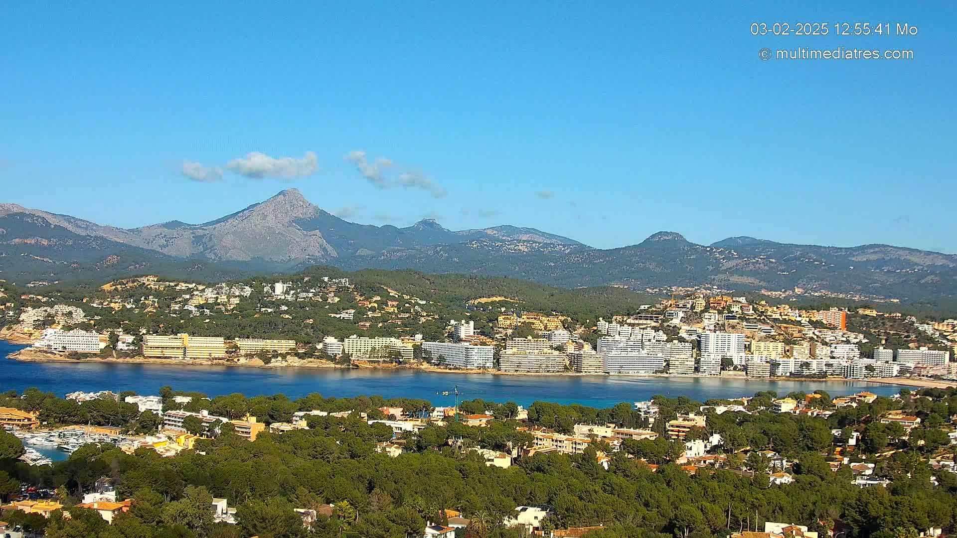 A coastal town nestled amongst green hills and mountains under a clear blue sky.