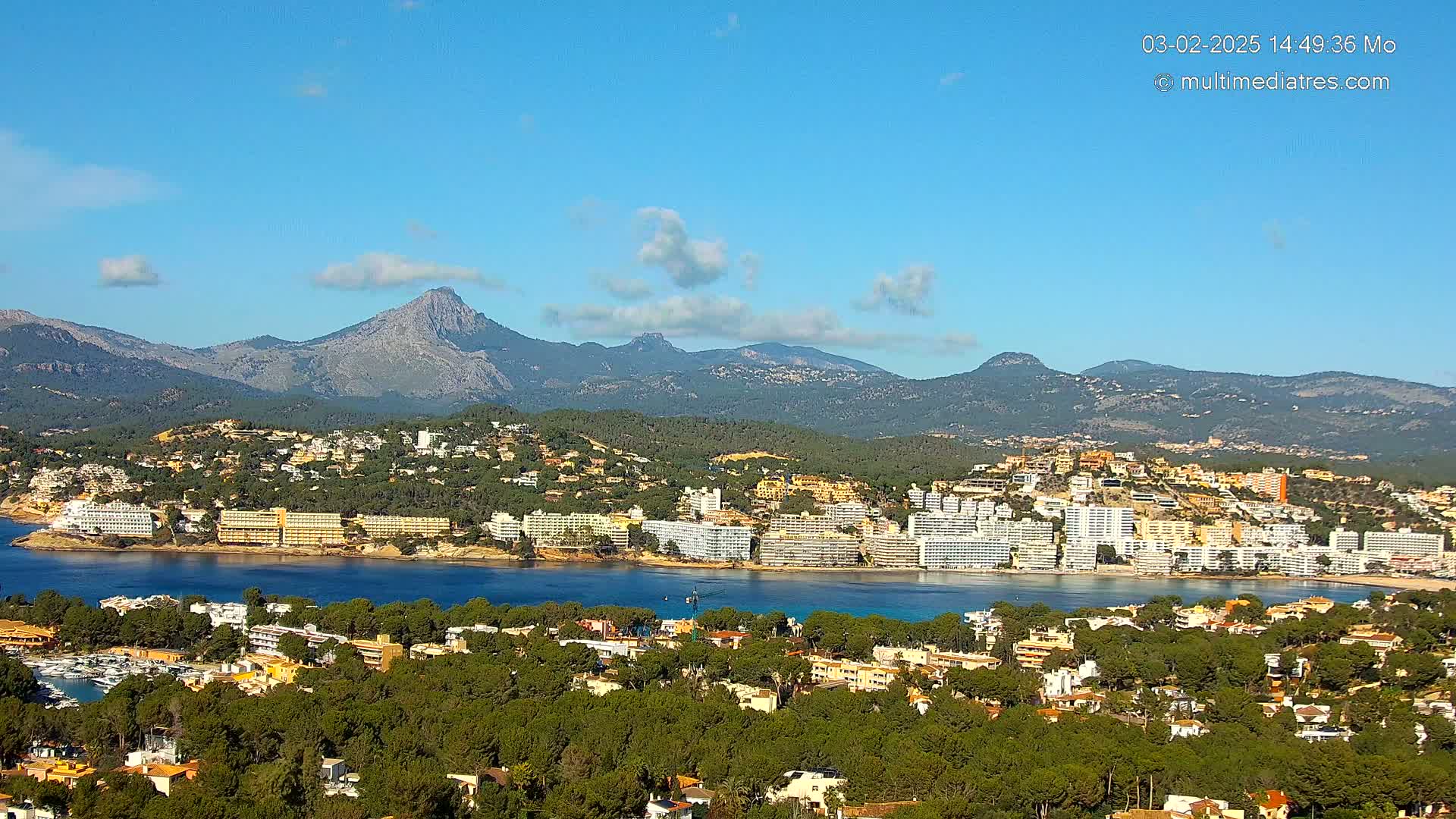 A coastal town nestled among green hills and mountains under a mostly clear blue sky.