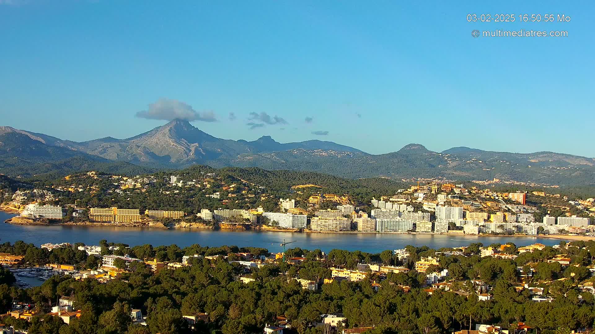 A coastal town with hotels and dense vegetation nestled against a mountain range under a clear blue sky.