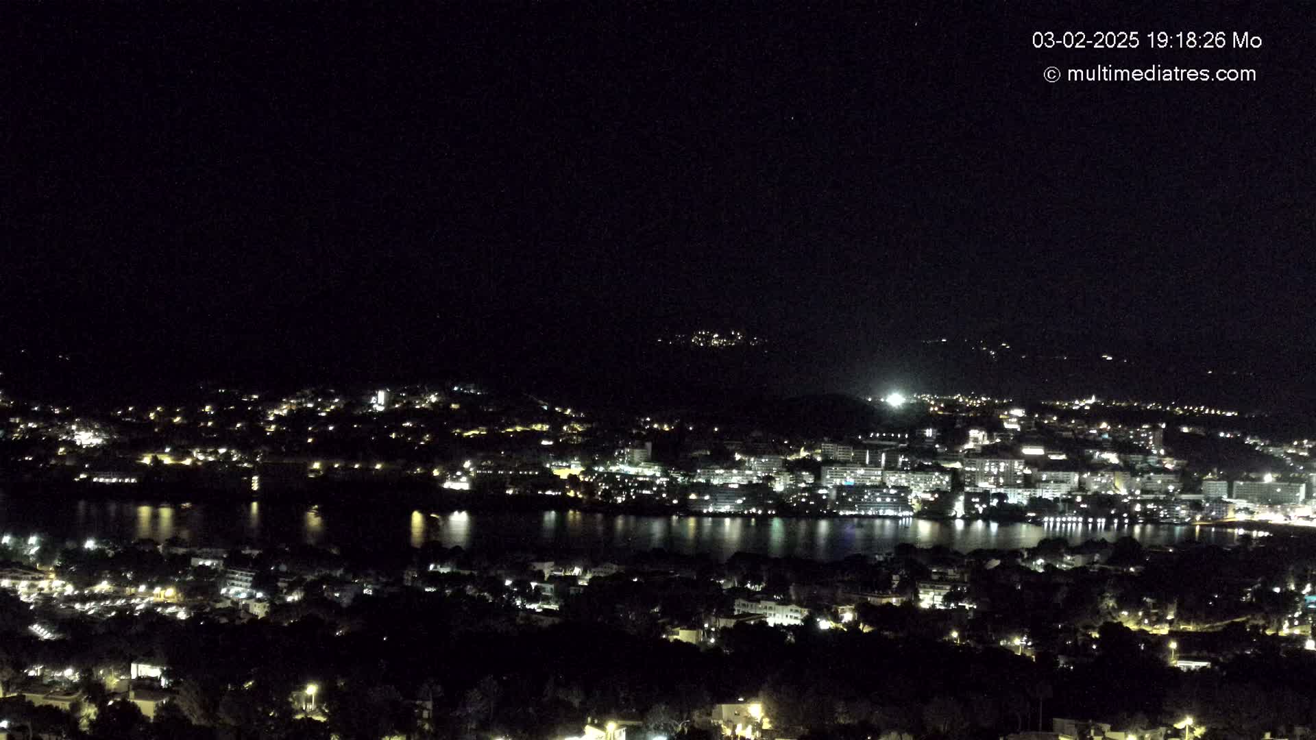 A nighttime, high-angle view of a city situated beside a body of water, with scattered lights illuminating buildings and surrounding areas under a clear, dark sky.