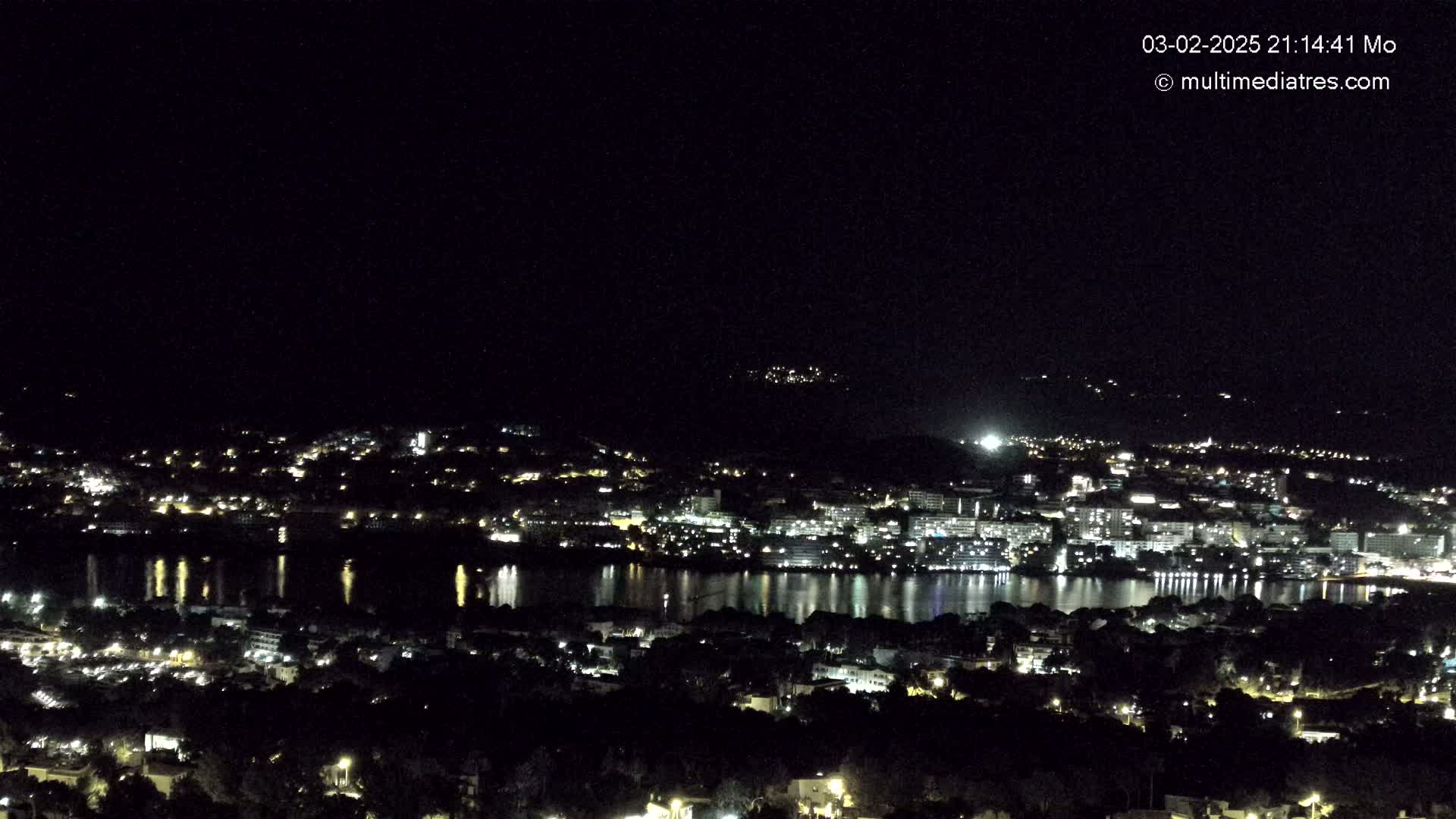 A nighttime, high-angle view of a city illuminated by lights surrounding a body of water reflecting the lights.