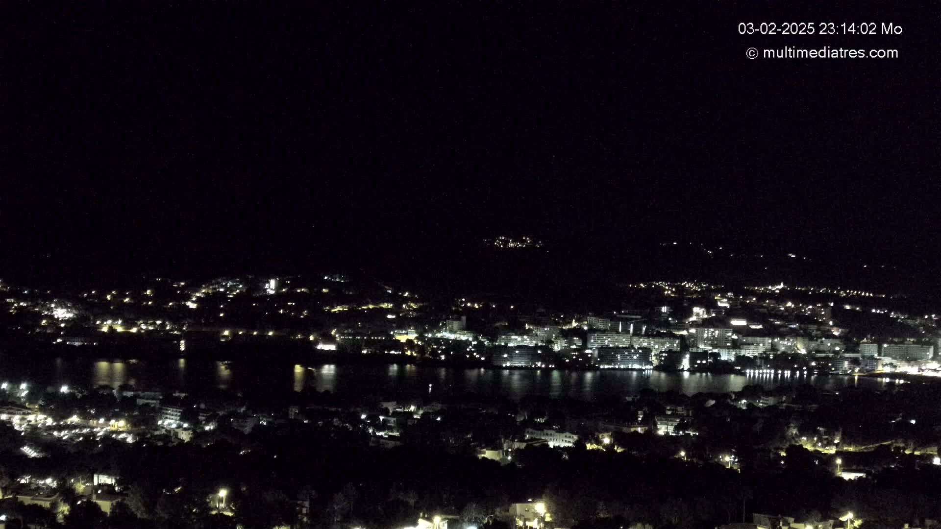 A nighttime, high-angle view shows a coastal town with its lights illuminating the waterfront and surrounding hills.