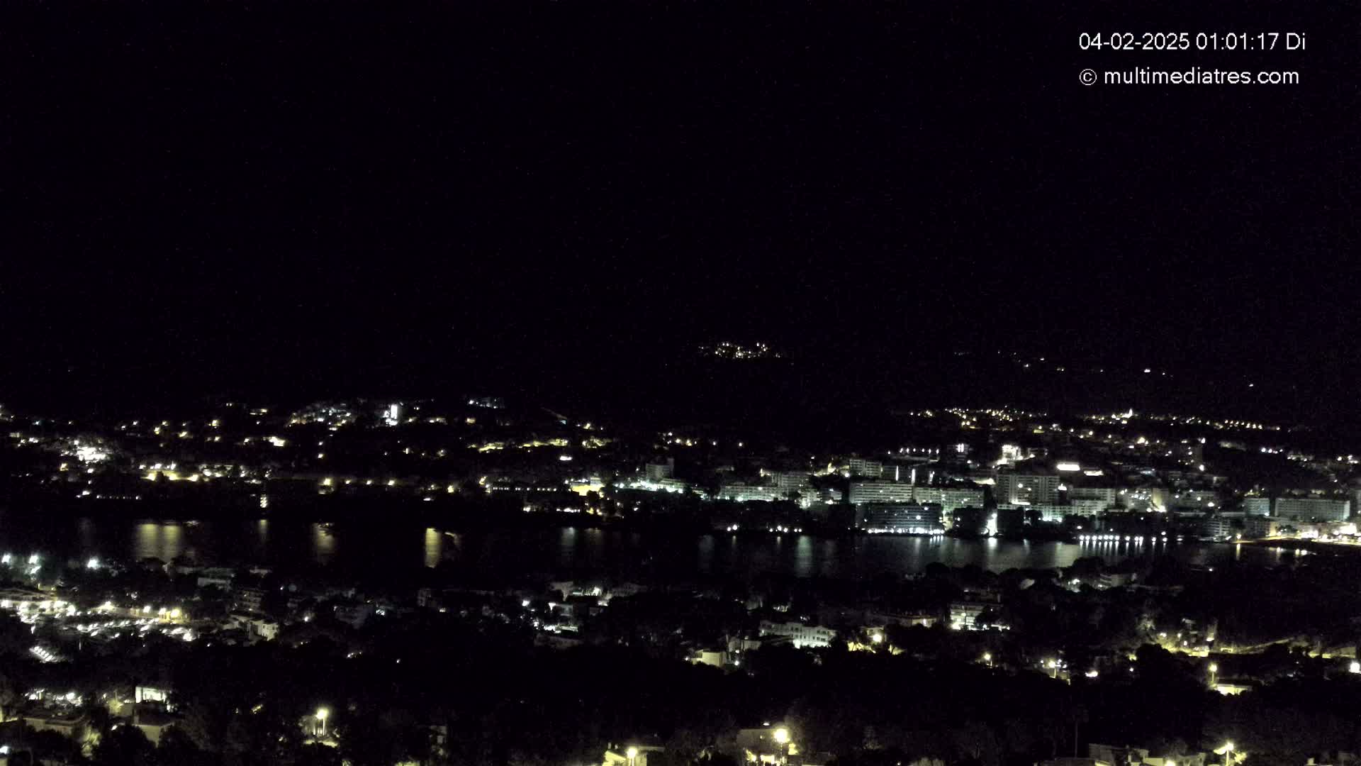 A nighttime view of a coastal town illuminated by lights, with a body of dark water reflecting the city lights under a clear, dark sky.
