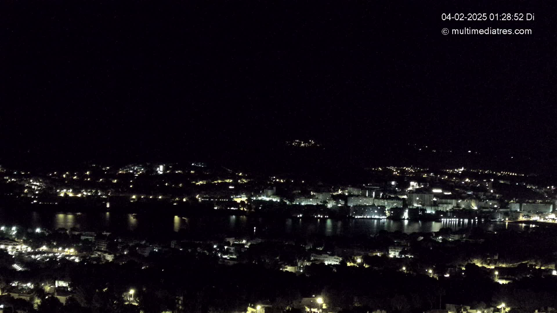 A nighttime, high-angle view shows a coastal town with its lights illuminating the buildings and streets, beside a dark body of water under a clear, starless sky.