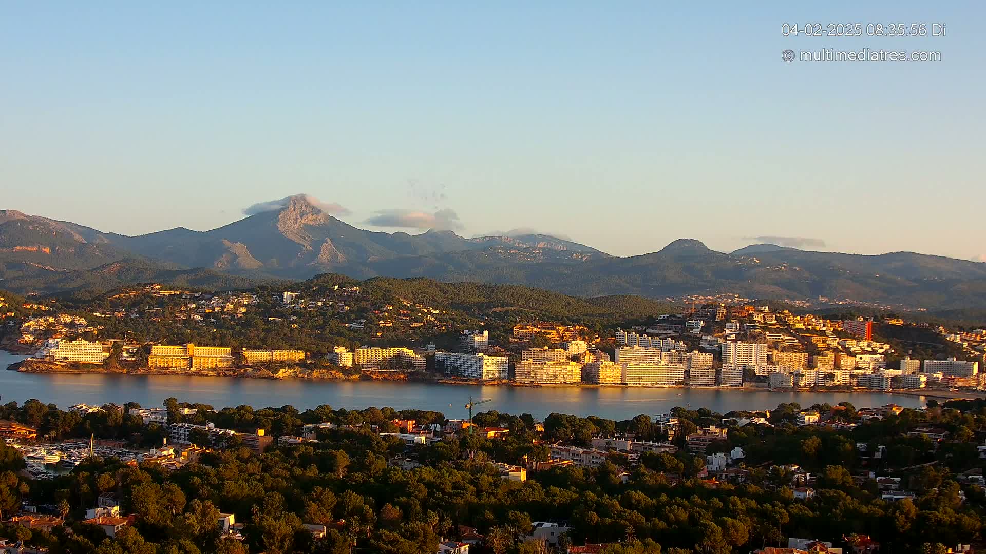 A coastal town with hotels and buildings nestled against a mountain range under a clear, light blue sky.