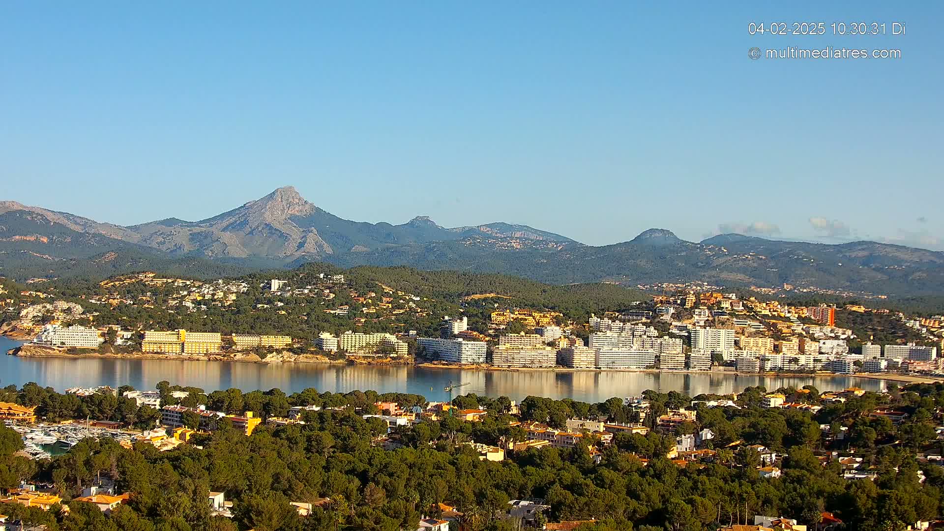 A coastal town nestled beside a calm bay, backed by a range of mountains under a clear blue sky.