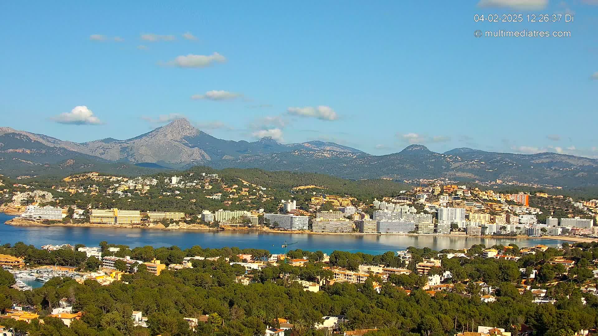 A coastal town with numerous buildings nestled between a calm bay and a mountain range under a mostly sunny sky.