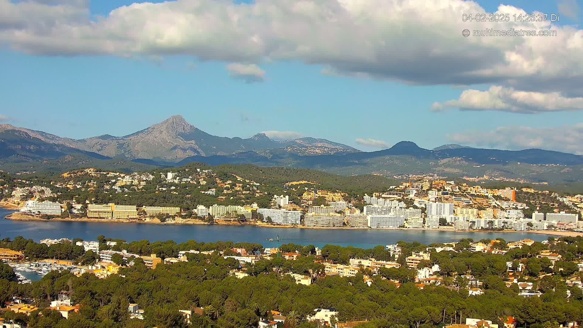 A coastal town nestled among green hills and mountains under a partly cloudy sky.