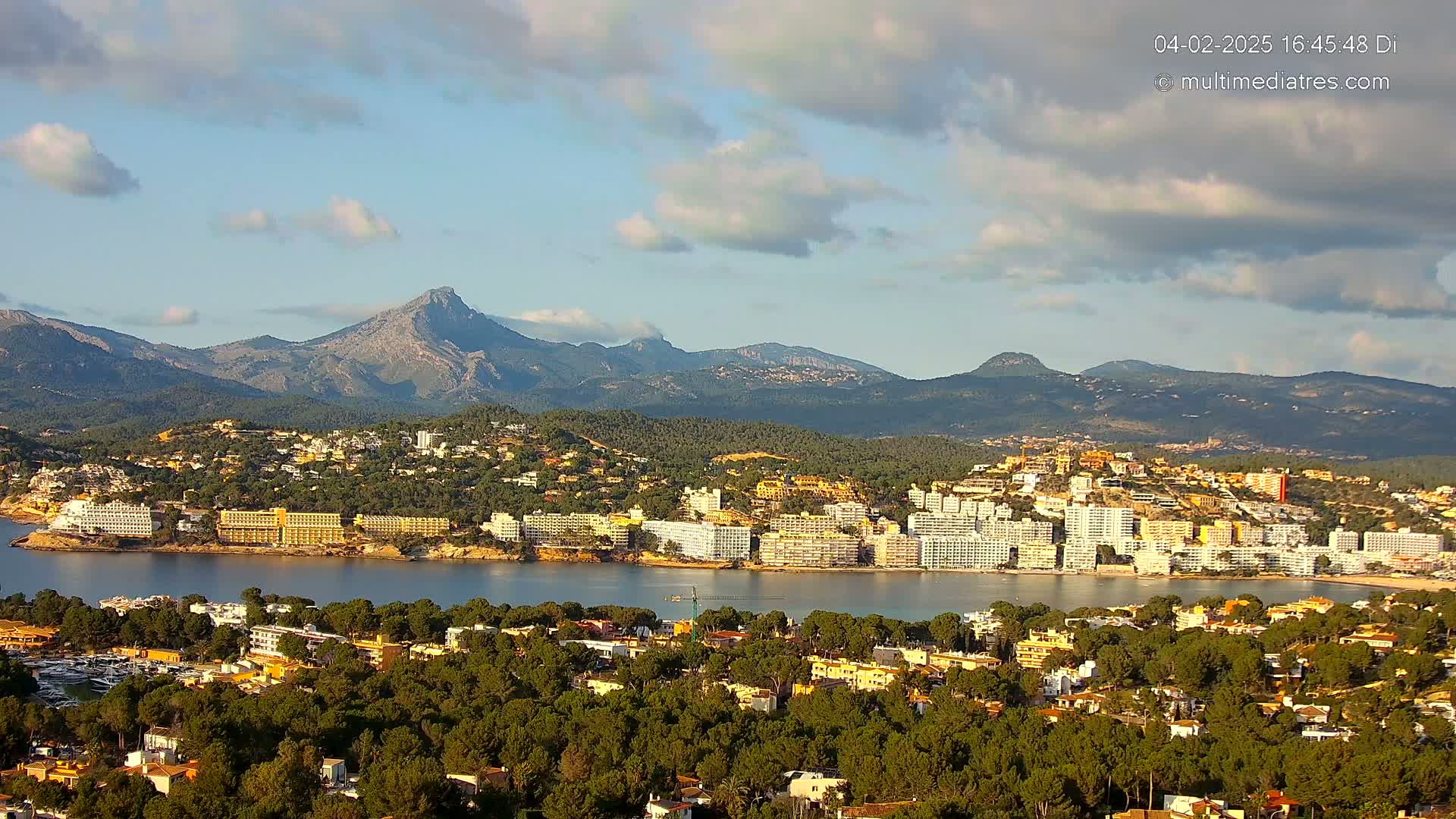 A coastal town with numerous hotels and buildings sits nestled between a calm bay and a range of mountains under a partly cloudy sky.
