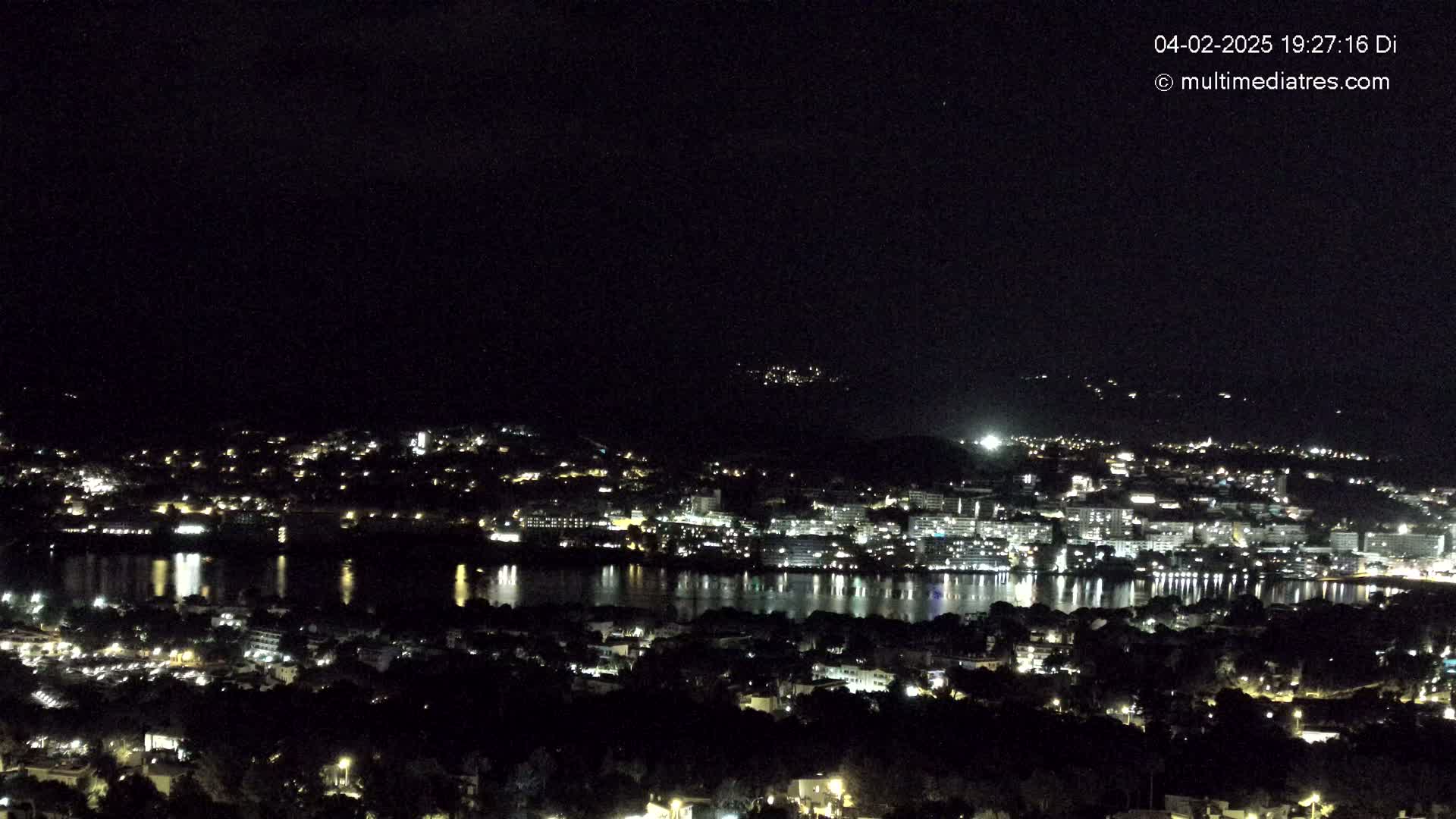 A nighttime, high-angle view shows a city illuminated by lights surrounding a dark body of water reflecting the city lights, under a clear, dark sky.