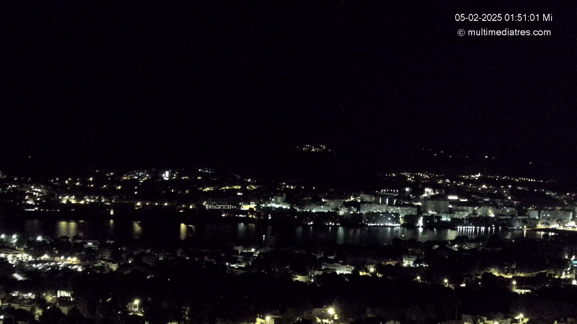 A nighttime, high-angle view shows a coastal town with its lights illuminating the buildings and a body of dark water.