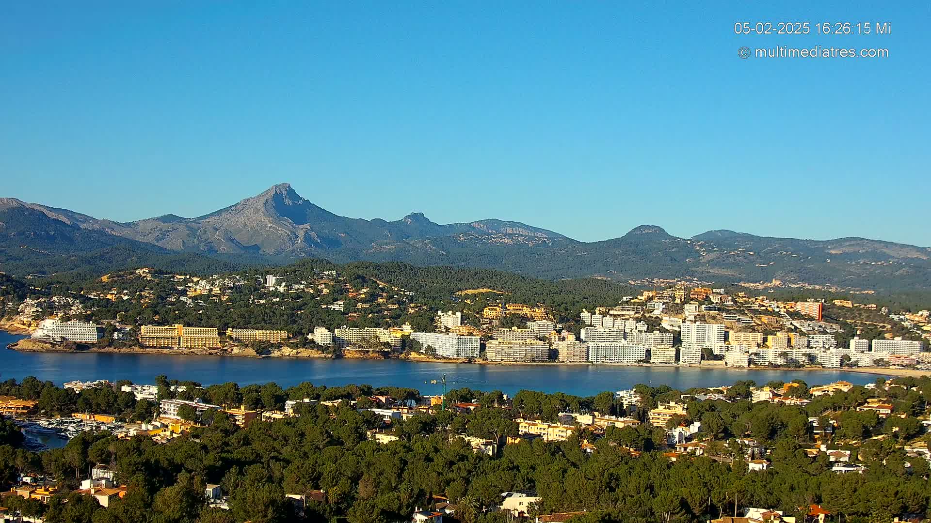 A coastal town nestled between a calm bay and a mountain range under a clear blue sky.