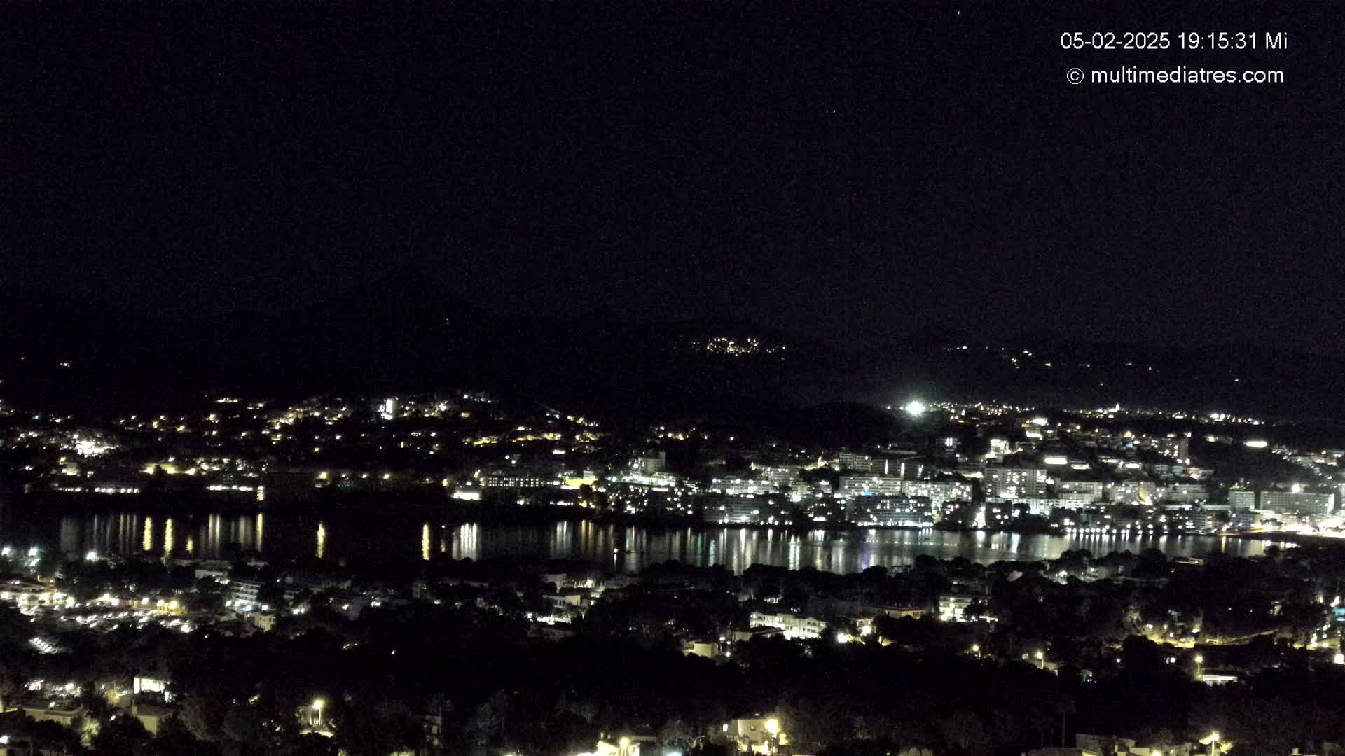 A nighttime view of a city illuminated by lights beside a body of water, reflecting the lights, with dark hills in the background under a clear, dark sky.