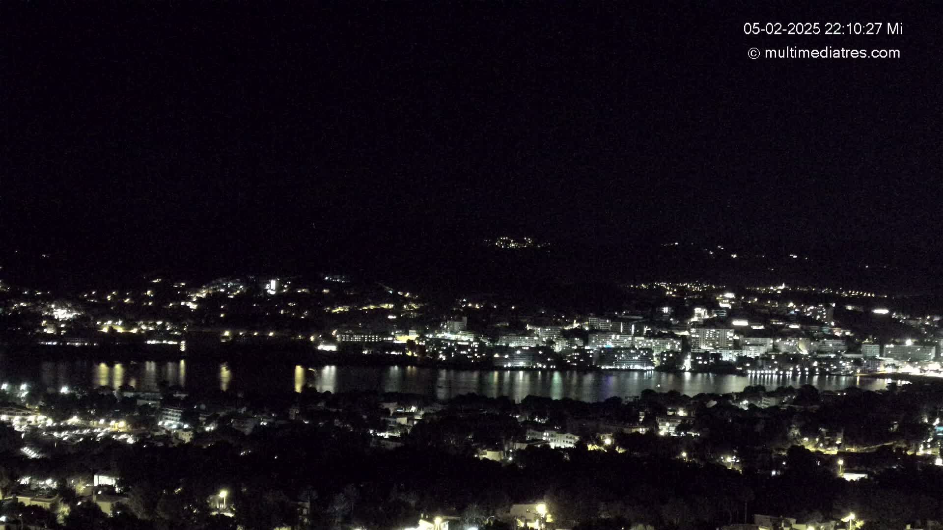 A nighttime view of a city illuminated by lights, situated beside a body of dark water reflecting the city's lights.