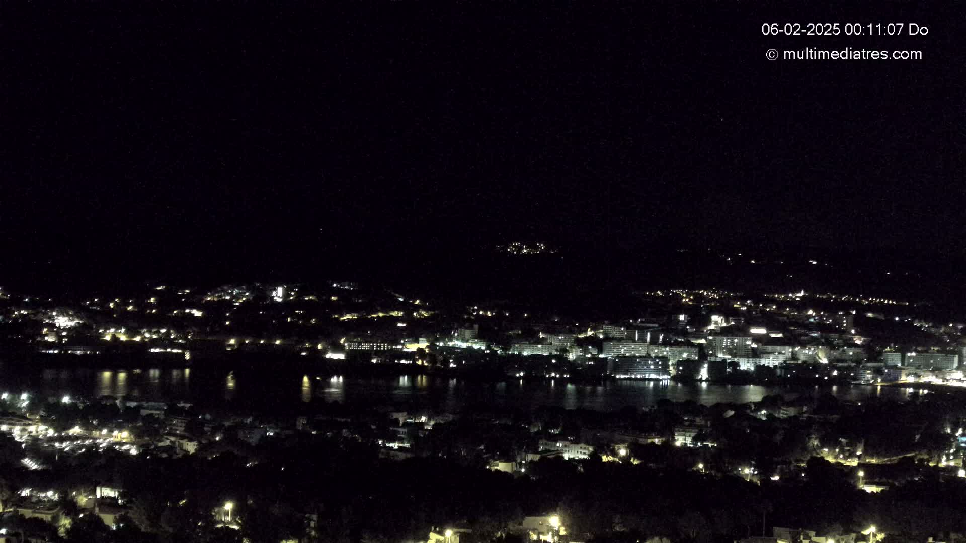 A nighttime view of a city spread across a body of water, with lights illuminating buildings and surrounding areas under a clear, dark sky.