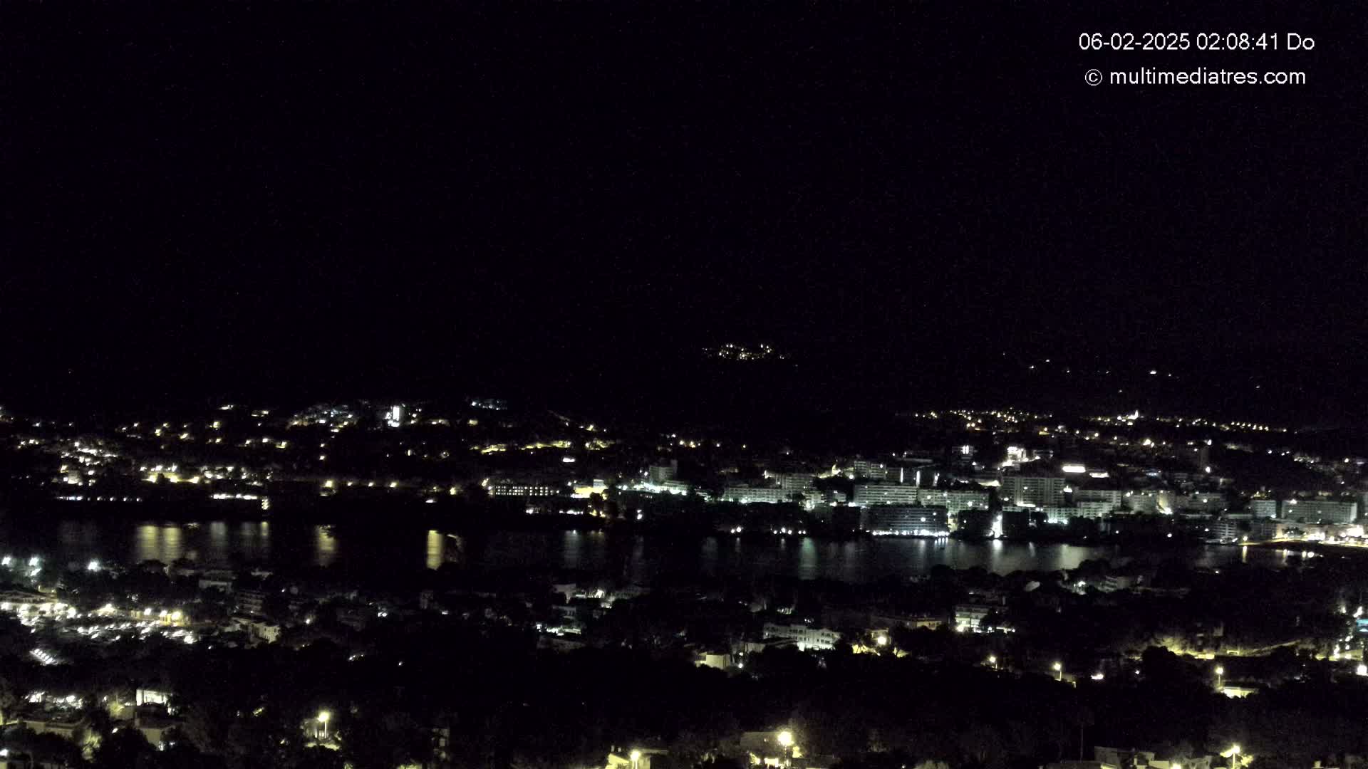 A nighttime, high-angle view shows a city with lights spread across the landscape, beside a body of dark water reflecting the city lights, under a clear, dark sky.