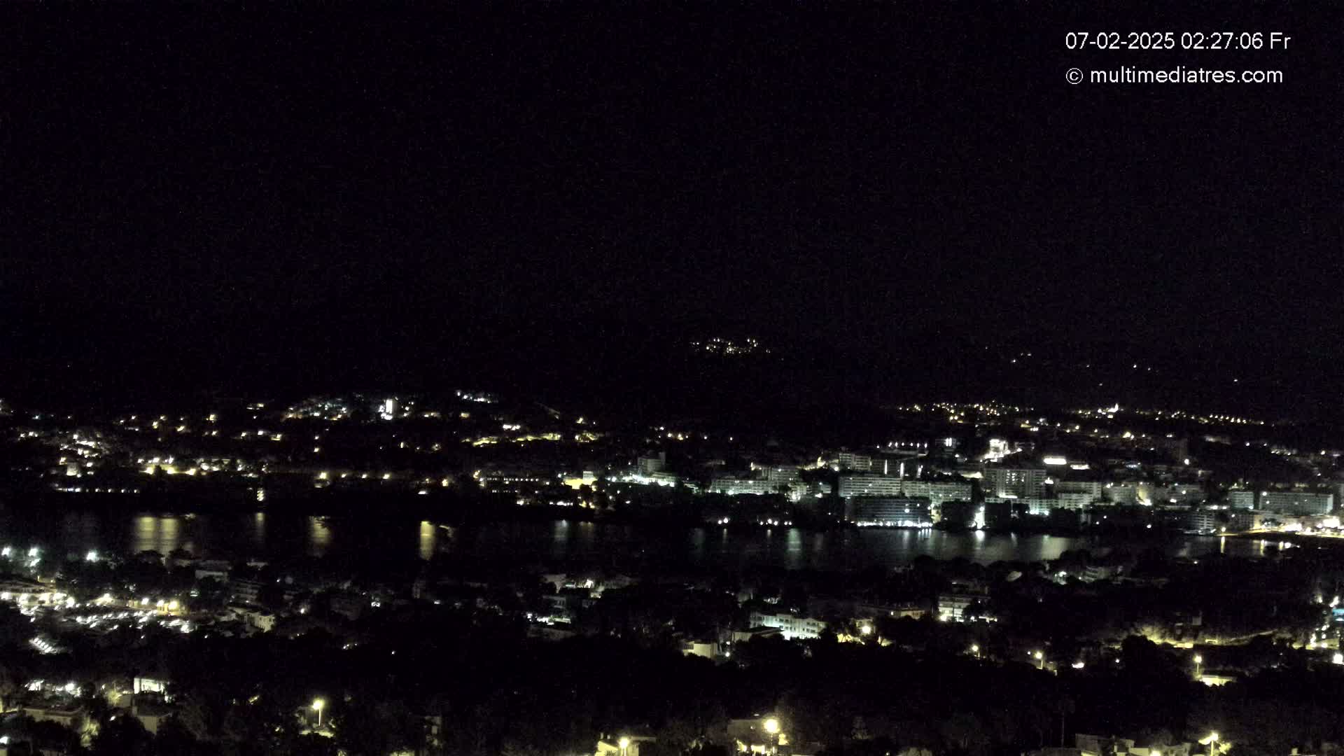 A nighttime, high-angle view of a city situated around a body of water, with hills visible in the background under a clear, dark sky.