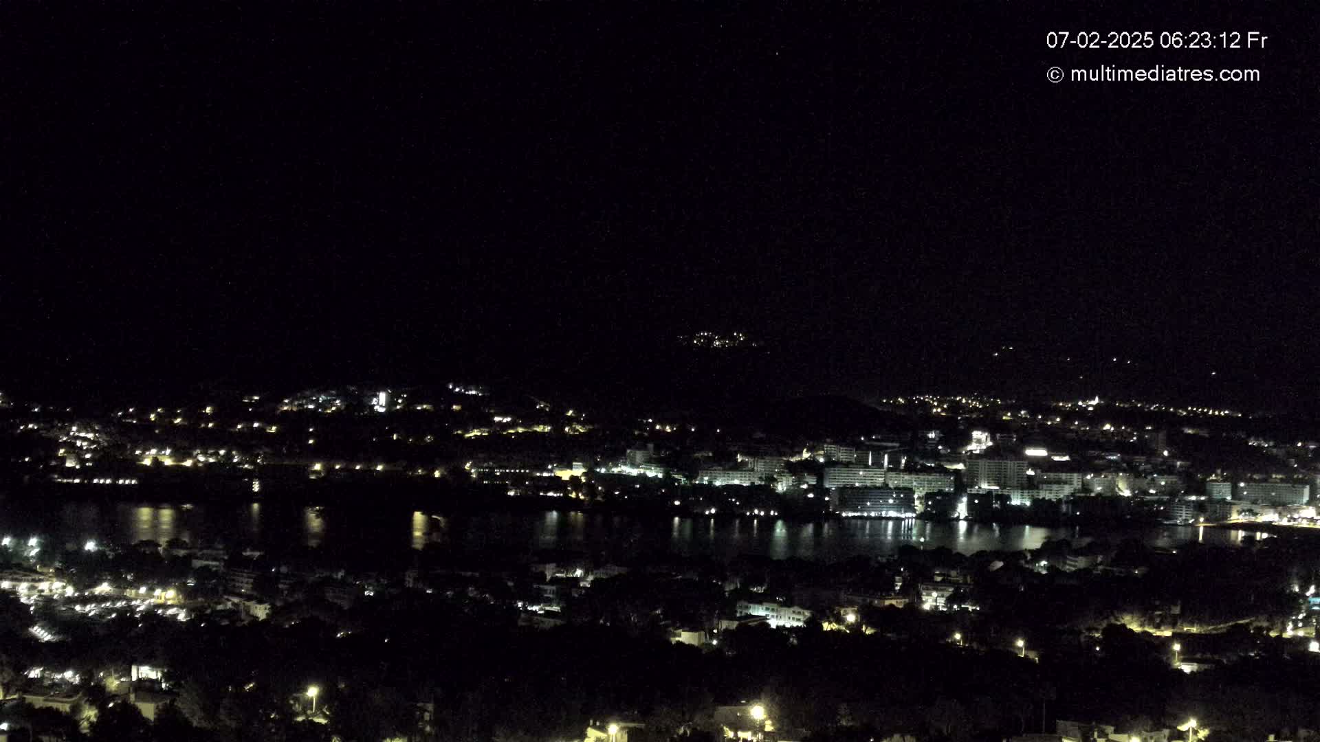 A nighttime, high-angle view of a city illuminated by lights, situated beside a body of dark water, with hills visible in the background under a clear, dark sky.
