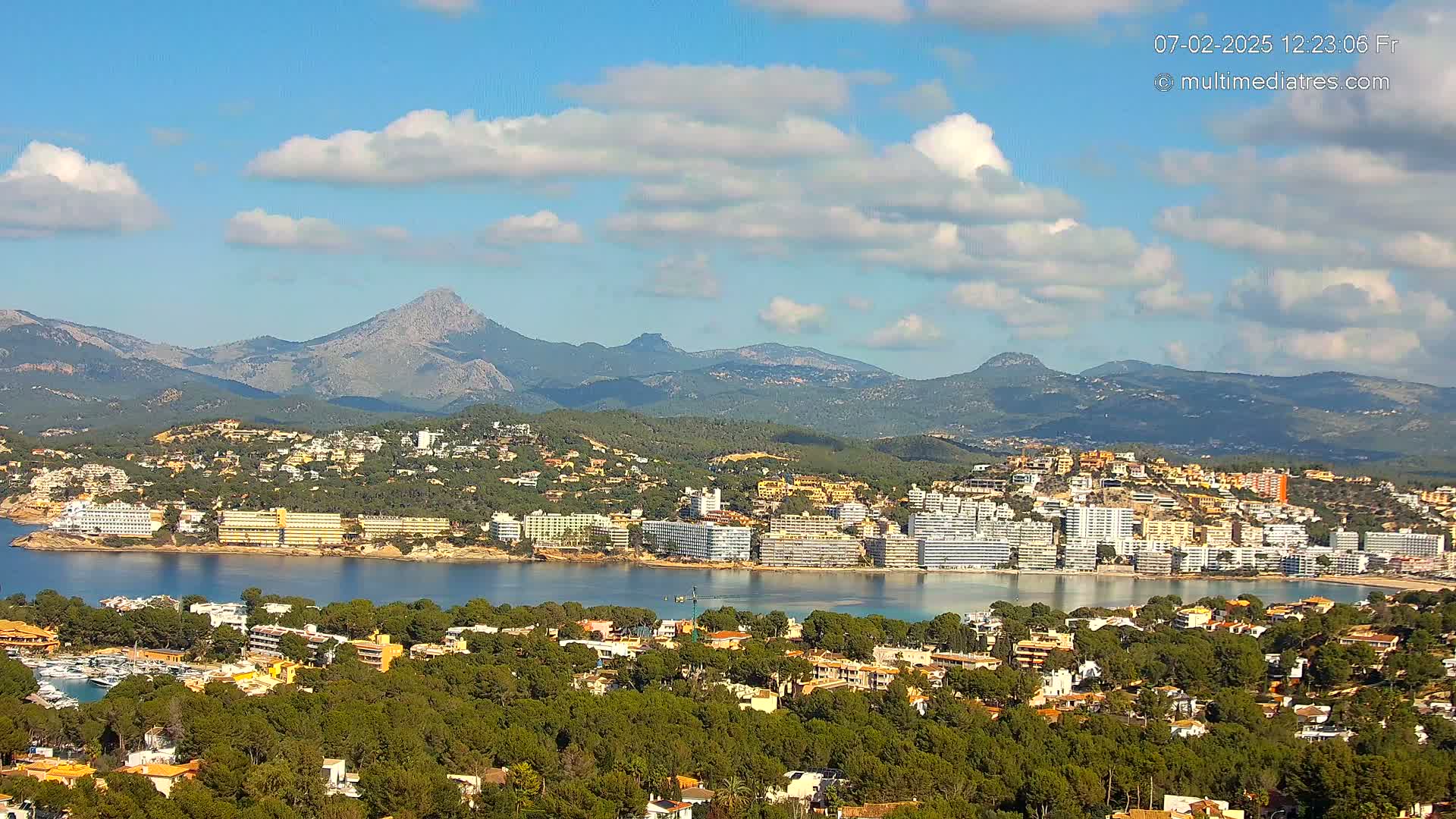 A coastal town with many buildings is nestled between a calm bay and a mountain range under a partly cloudy sky.