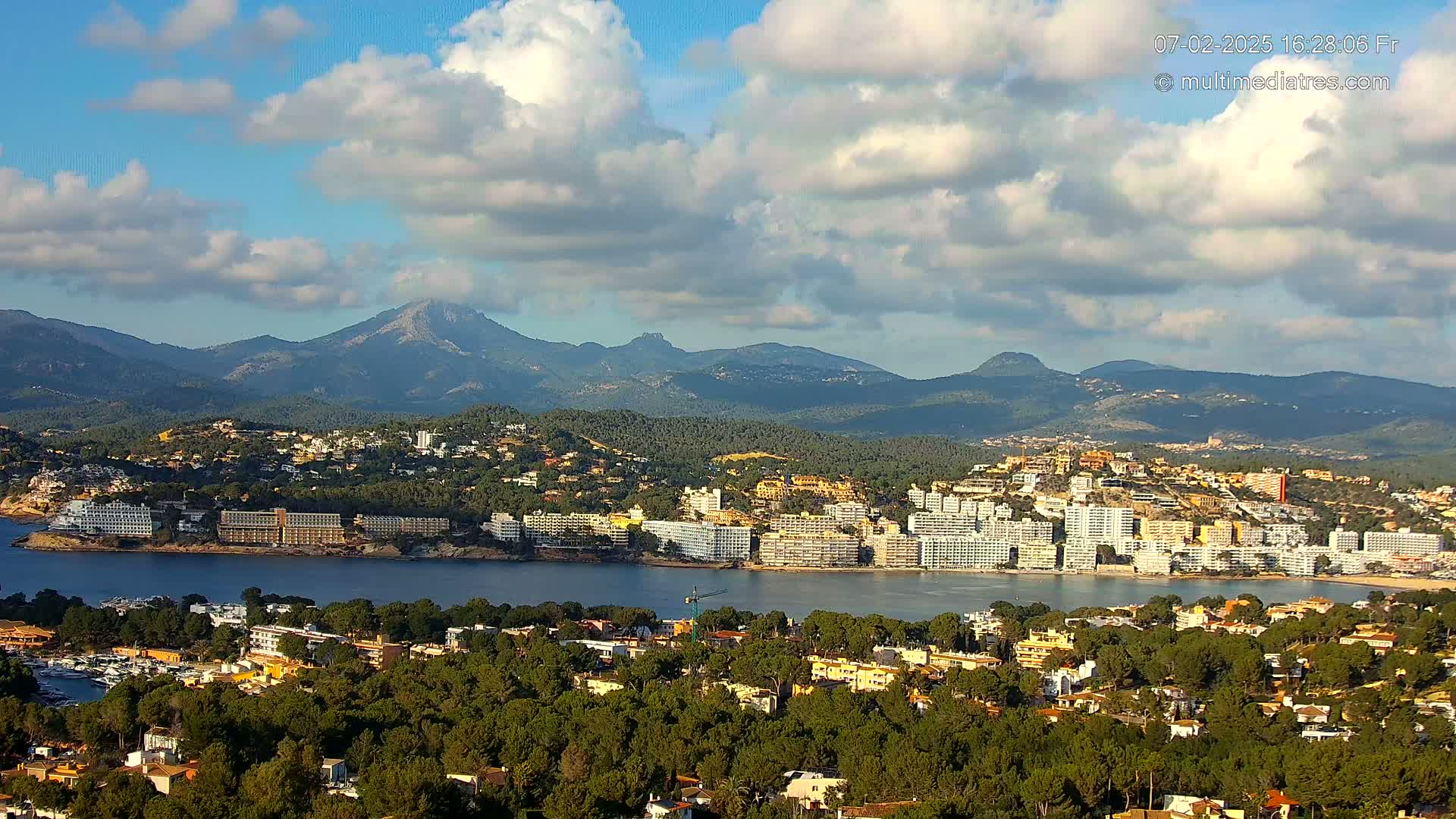 A coastal town with numerous buildings, nestled among green hills and backed by a mountain range under a partly cloudy sky.