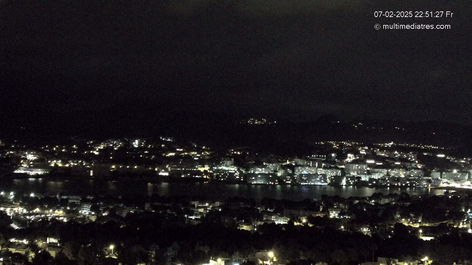 A nighttime view of a coastal city illuminated by lights, with a dark sky and calm water.